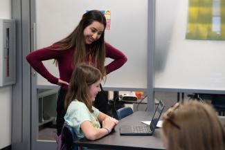 A classroom teacher instructs a young student on a laptop